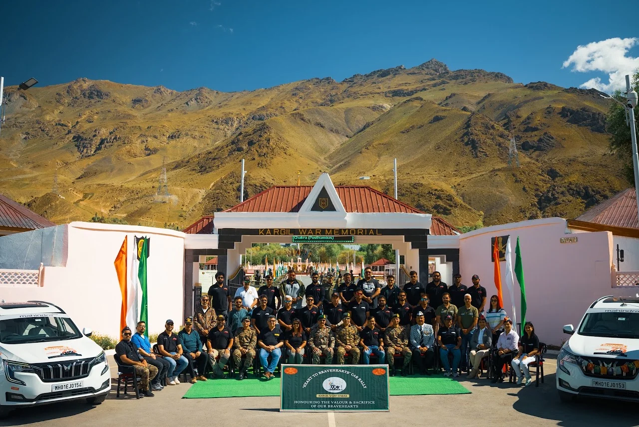 People at the Kargil War Memorial in Dras, with Tololing Peak behind them and two Mahindra SUVs nearby