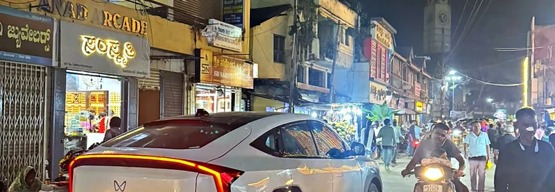A busy night street in Dharwad with lit Kannada shop signboards and a white Mahindra XEV 9e in the foreground