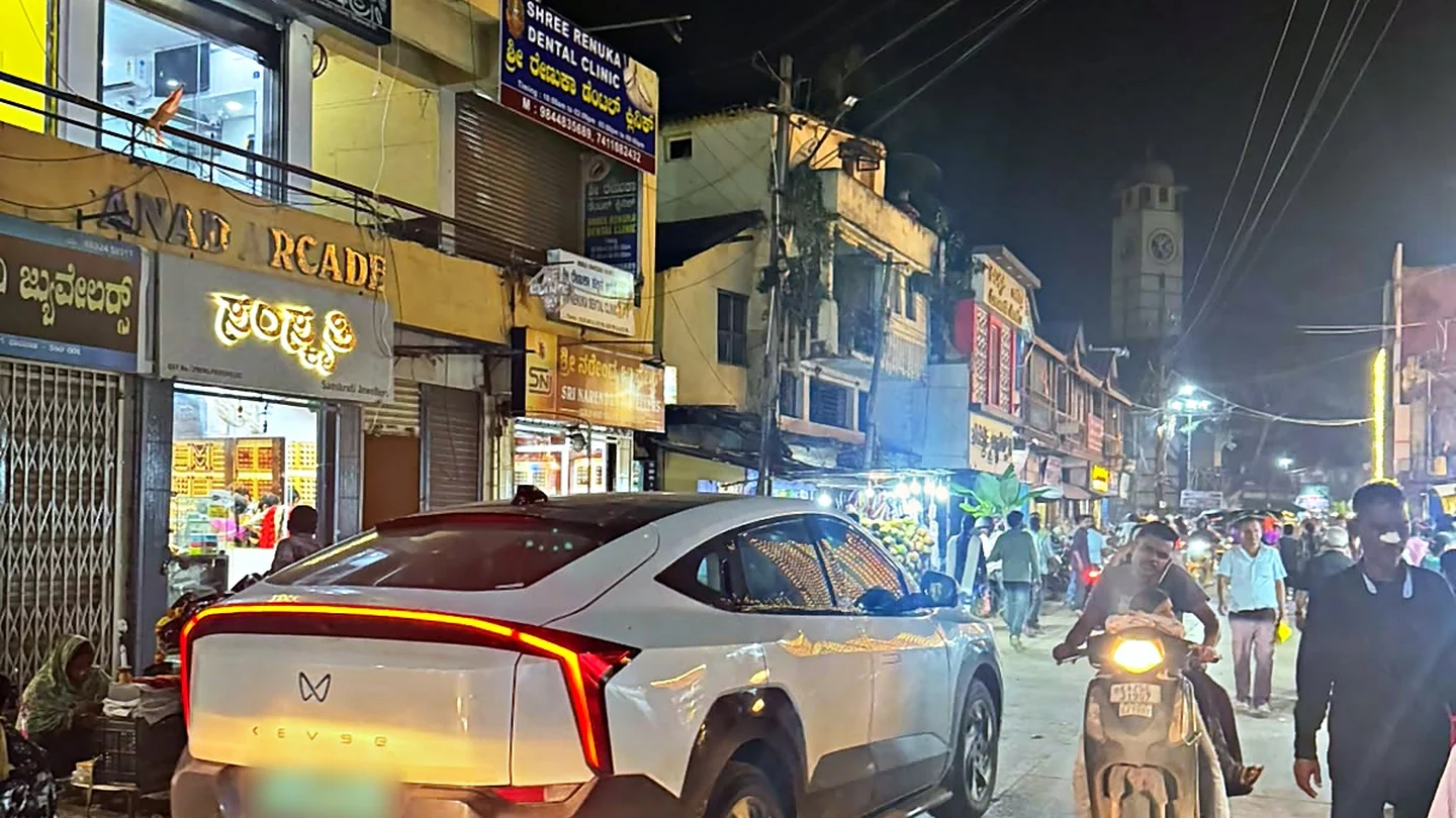 A busy night street in Dharwad with lit Kannada shop signboards and a white Mahindra XEV 9e in the foreground